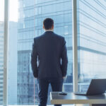 Back View Of The Thoughtful Businessman Wearing A Suit Standing In His Office, Hands In Pockets And Contemplating Next Big Business Deal, Looking Out Of The Window. Big City Panoramic Window View.